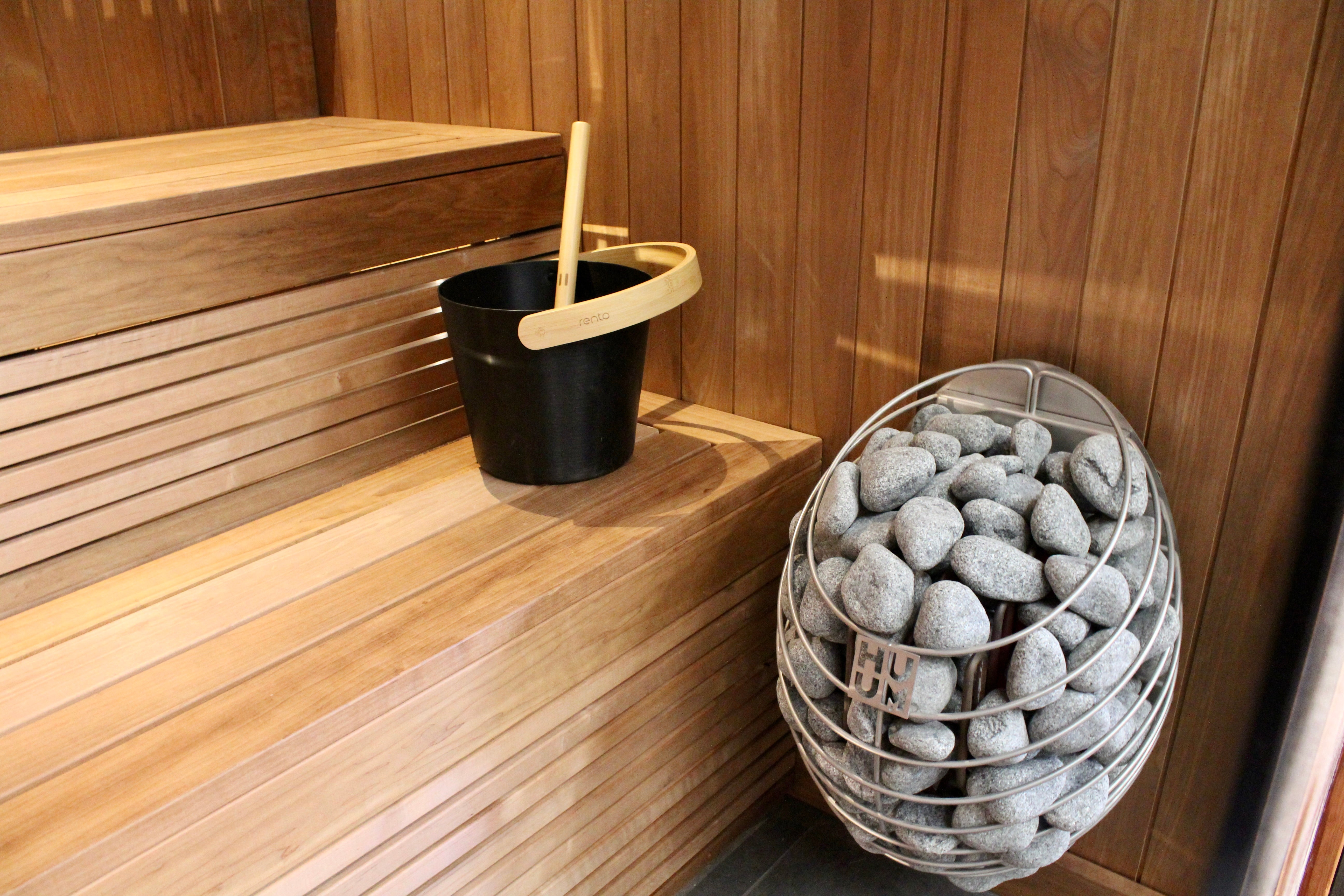 Black bucket with a wooden ladle on a wooden bench next to a basket of stones in a sauna.