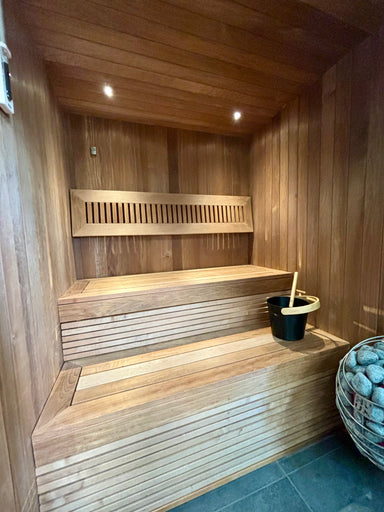 Wooden sauna with benches, a bucket, and a stone sauna heater on a tiled floor.